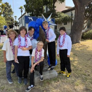 Group of boys in white shirts and purple leis posing around a dolphin statue with blue balloons at an outdoor school event.