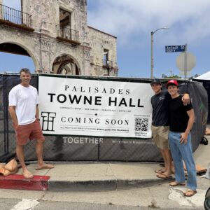 Three men stand in front of a construction fence and a banner reading 'PALISADES TOWNE HALL' beside a weathered ruins backdrop.