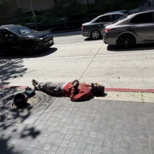 Man lying on the sidewalk beside a parked black SUV and a gray car, wearing red top and gray pants with a helmet nearby.