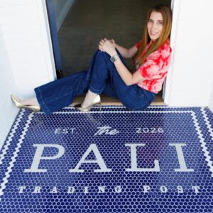 Smiling woman in jeans and a red tie-dye shirt sits cross-legged in a doorway on a blue hexagonal-tile mat that reads 'Est. 2016 PAL T Trading Post'.