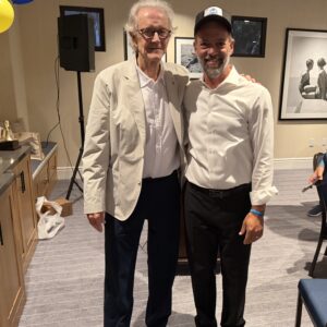 Two men pose together indoors at a casual event; older man with white hair and glasses, wearing a light blazer, beside a smiling man in a white shirt and cap with balloons and photos in the background responsibly.