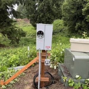 Outdoor weatherproof electrical disconnect box on a wooden post in a garden, with cables coiled at the base and surrounding greenery nearby