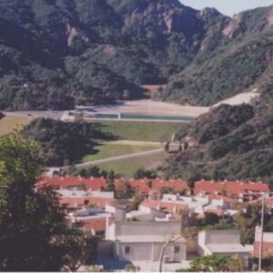 Hilly town with white houses and red-tiled roofs at the valley floor, mountains in the background and a distant airstrip or runway.