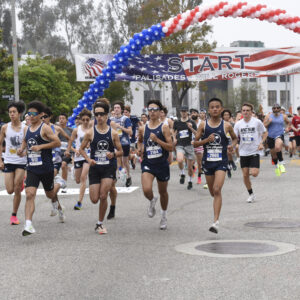 Runners at the start of a road race under a red, white, and blue balloon arch and START banner.