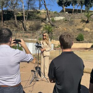 Female spokesperson speaks to reporters outdoors, microphones and cameras aimed at her, hillside in the background under a clear sky.