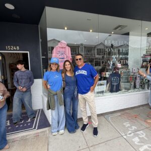 Group of smiling adults standing together outside a shop window, wearing blue shirts and jeans, posing for a photo