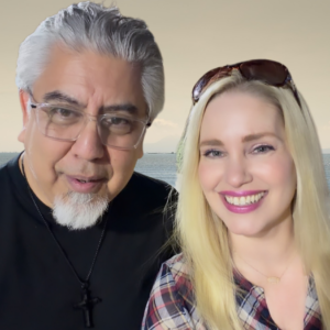 Two smiling people pose for a photo by the water: a man with gray hair and glasses wearing a black shirt and cross necklace, and a blonde woman in a plaid shirt with sunglasses on her head.